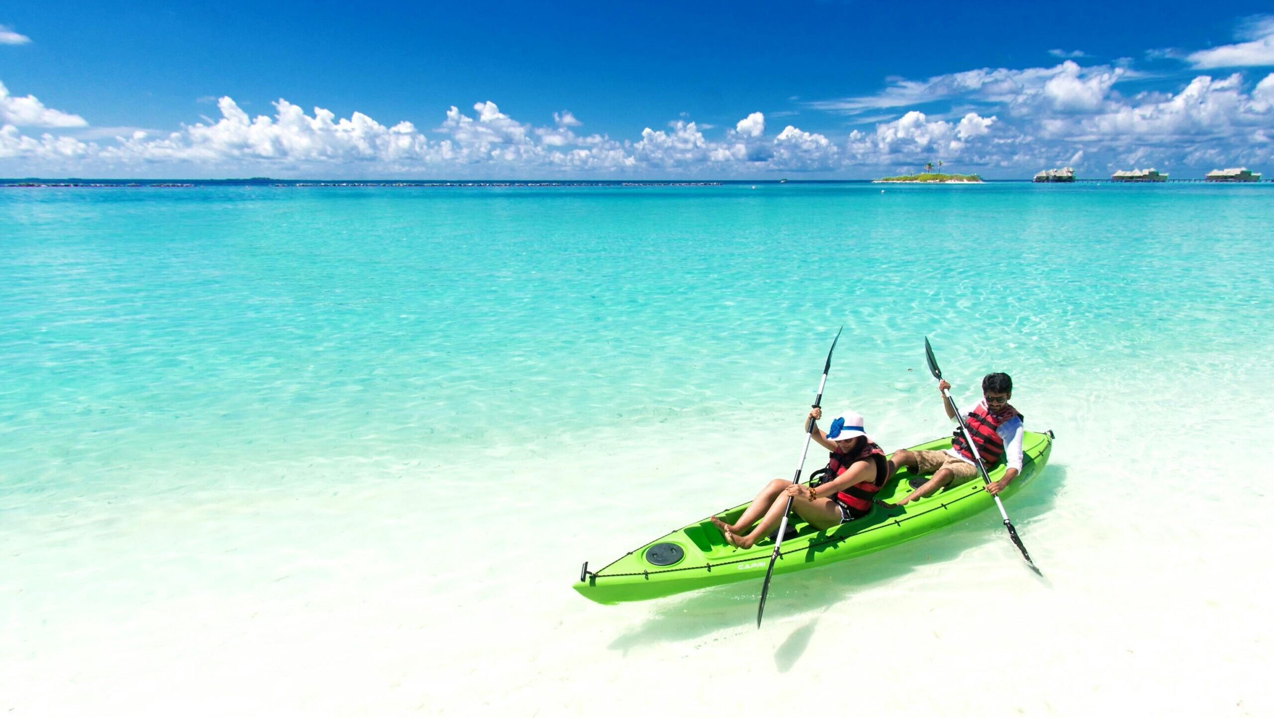 Two people kayaking in clear turquoise waters on a sunny tropical beach.