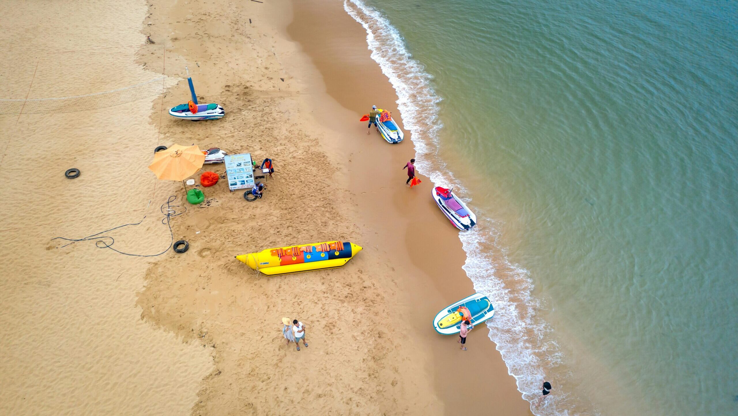 Drone shot of a sandy beach with people, boats, and a vibrant seashore, capturing vacation vibes.